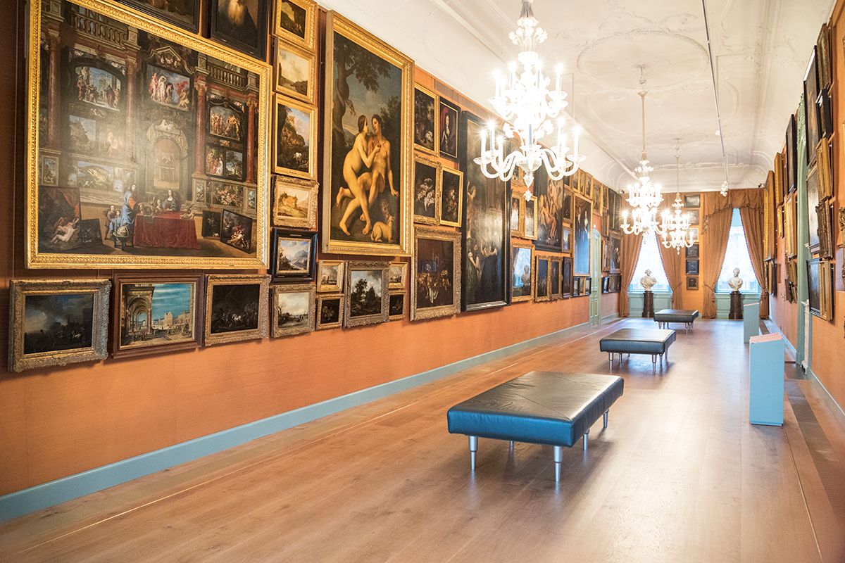 Een foto van de Galerij Prins Willem V. Je ziet een zaal met van top tot plint schilderijen, bankjes in het midden en kroonluchters aan het plafond.